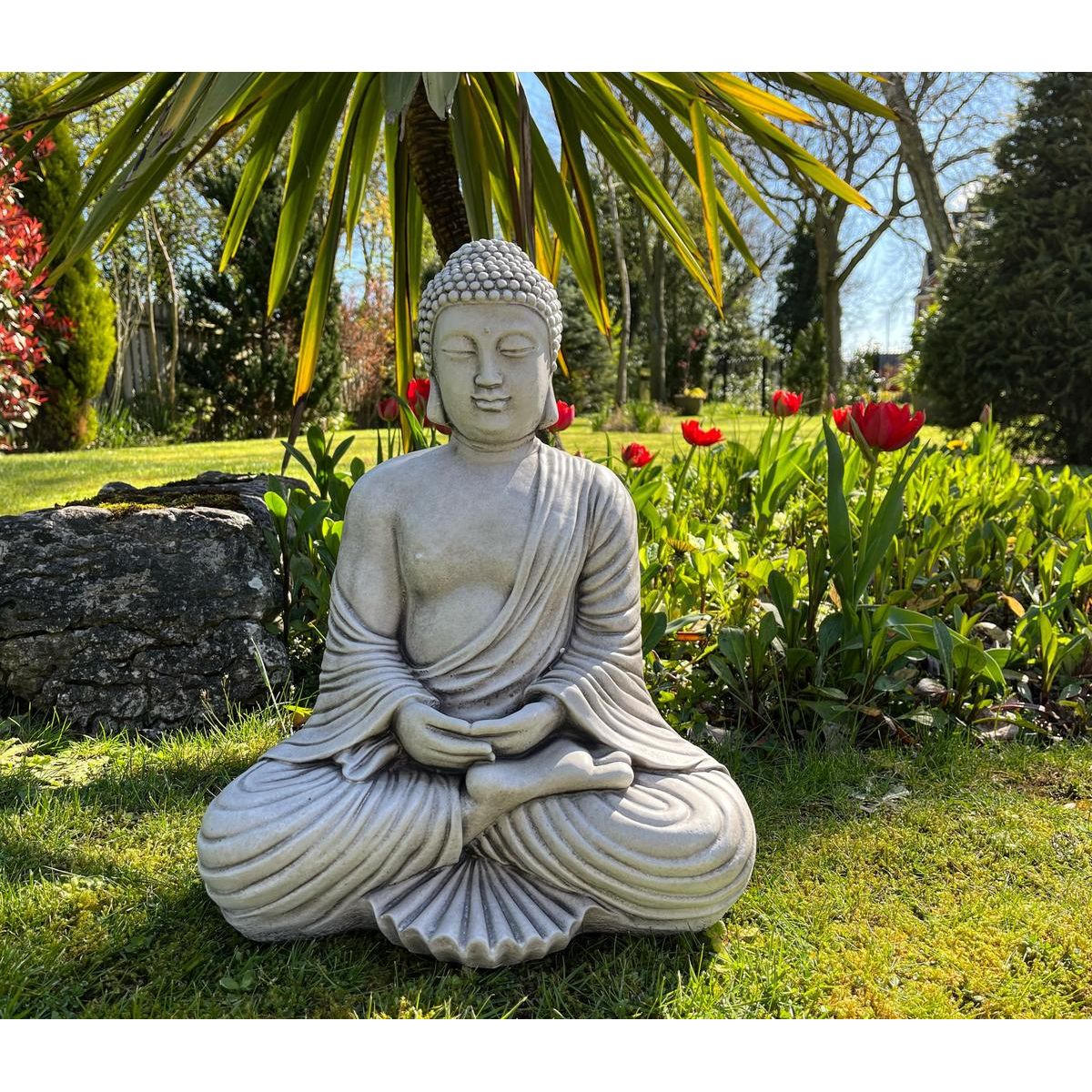 Stone Buddha statue in serene garden setting, surrounded by vibrant red tulips and lush greenery. Perfect for creating a peaceful outdoor meditation space.