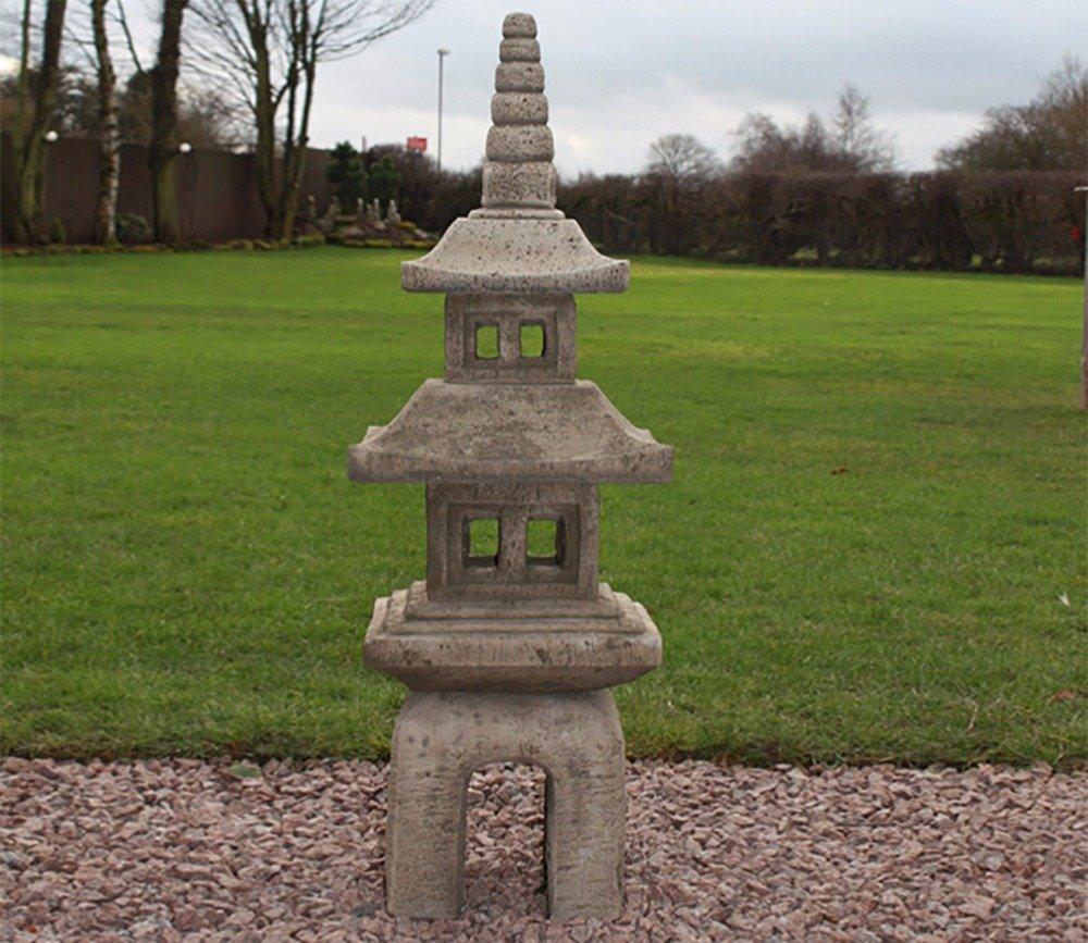 A grey stone Japanese pagoda lantern stands on gravel in a garden, featuring multiple tiers and square openings, set against a lush green lawn and trees.
