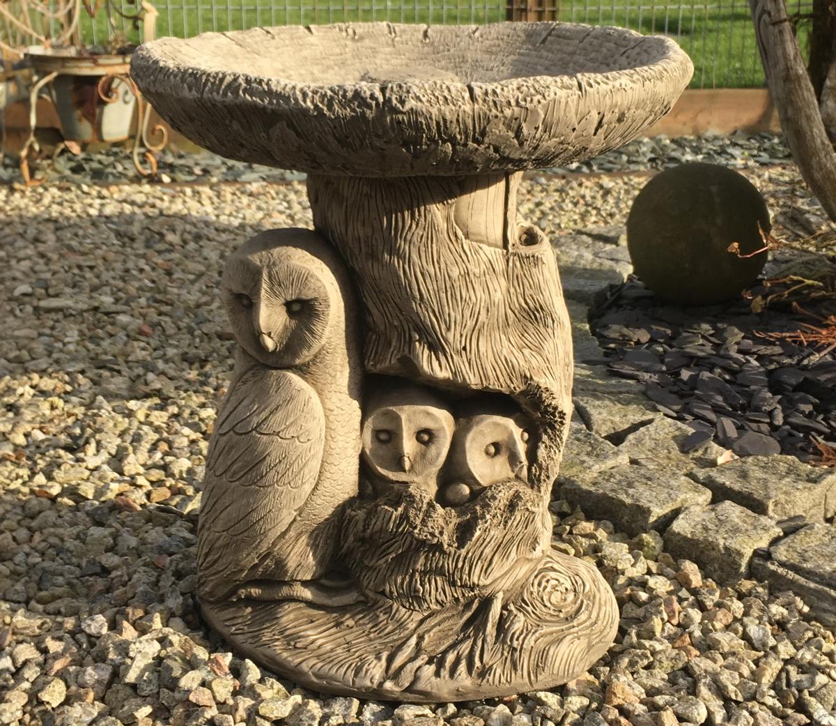Stone bird bath sculpture with owl family design, featuring a large owl and two smaller owls nestled in a tree trunk-like base, set on a gravel garden path.