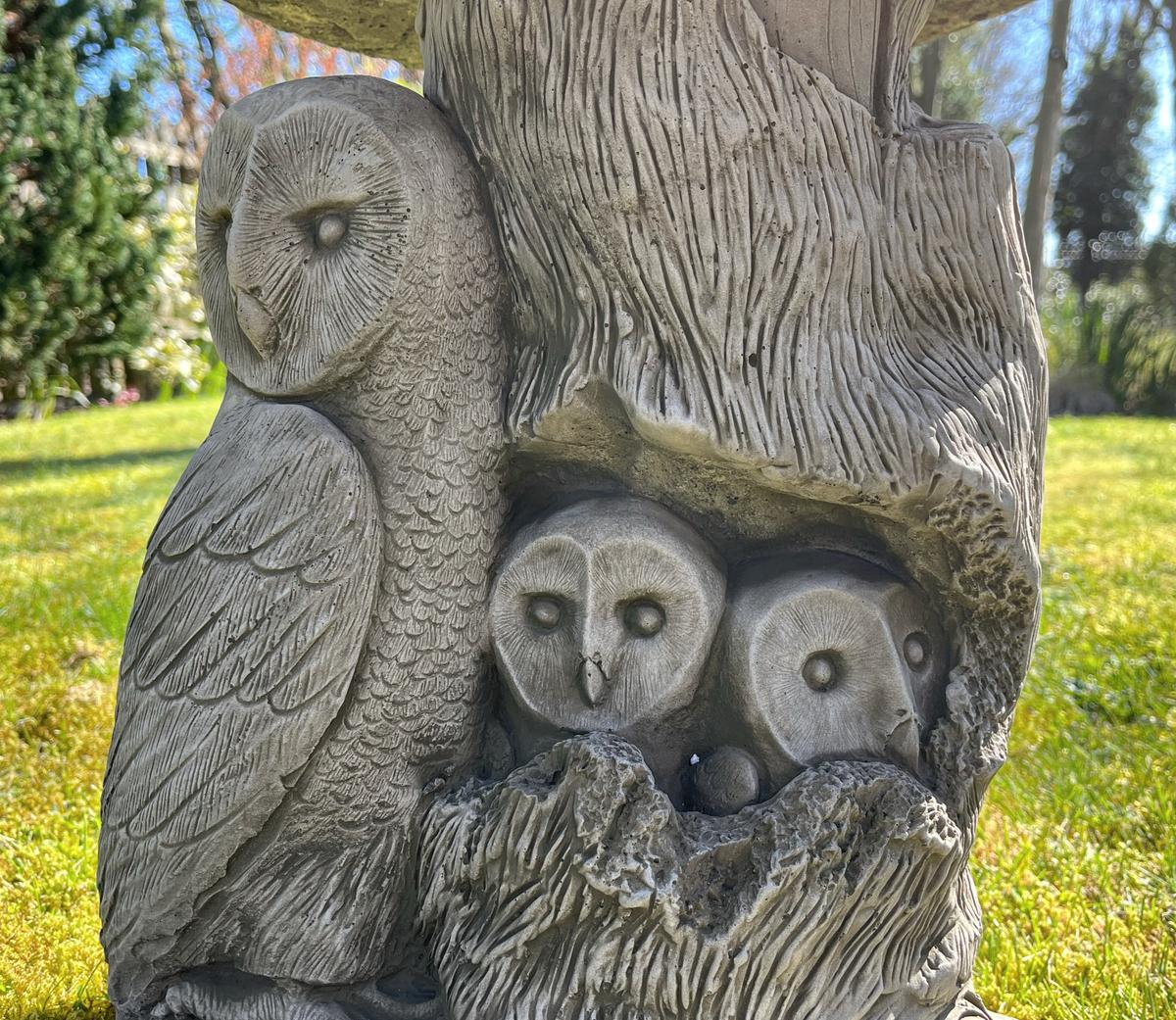 A detailed stone owl sculpture in a garden, featuring a large owl and two smaller owlets nestled in a tree trunk, highlighting intricate feather details.