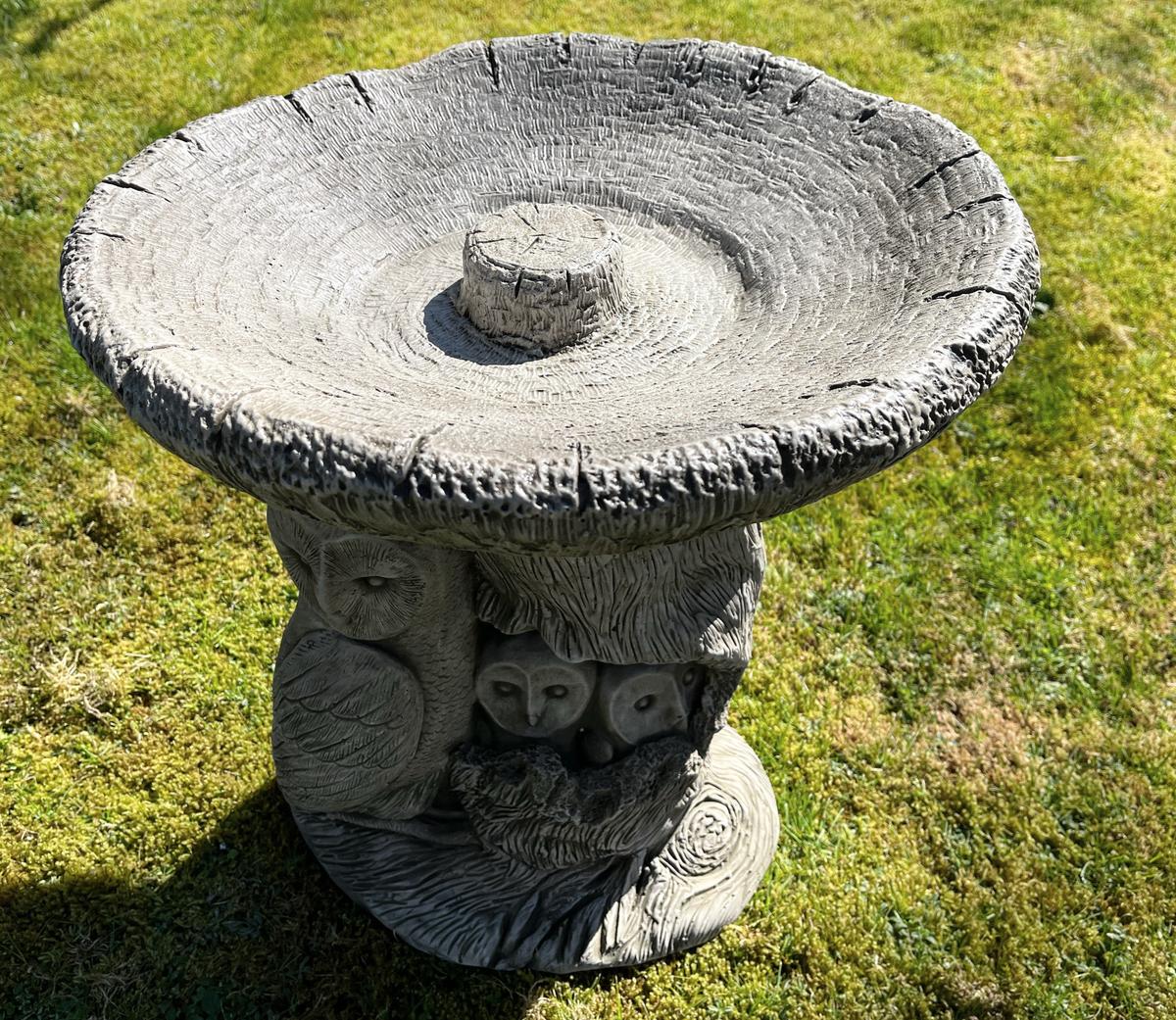 Stone bird bath with a textured circular top and intricate owl carvings on a grassy background. Ideal for enhancing garden decor and attracting wildlife.