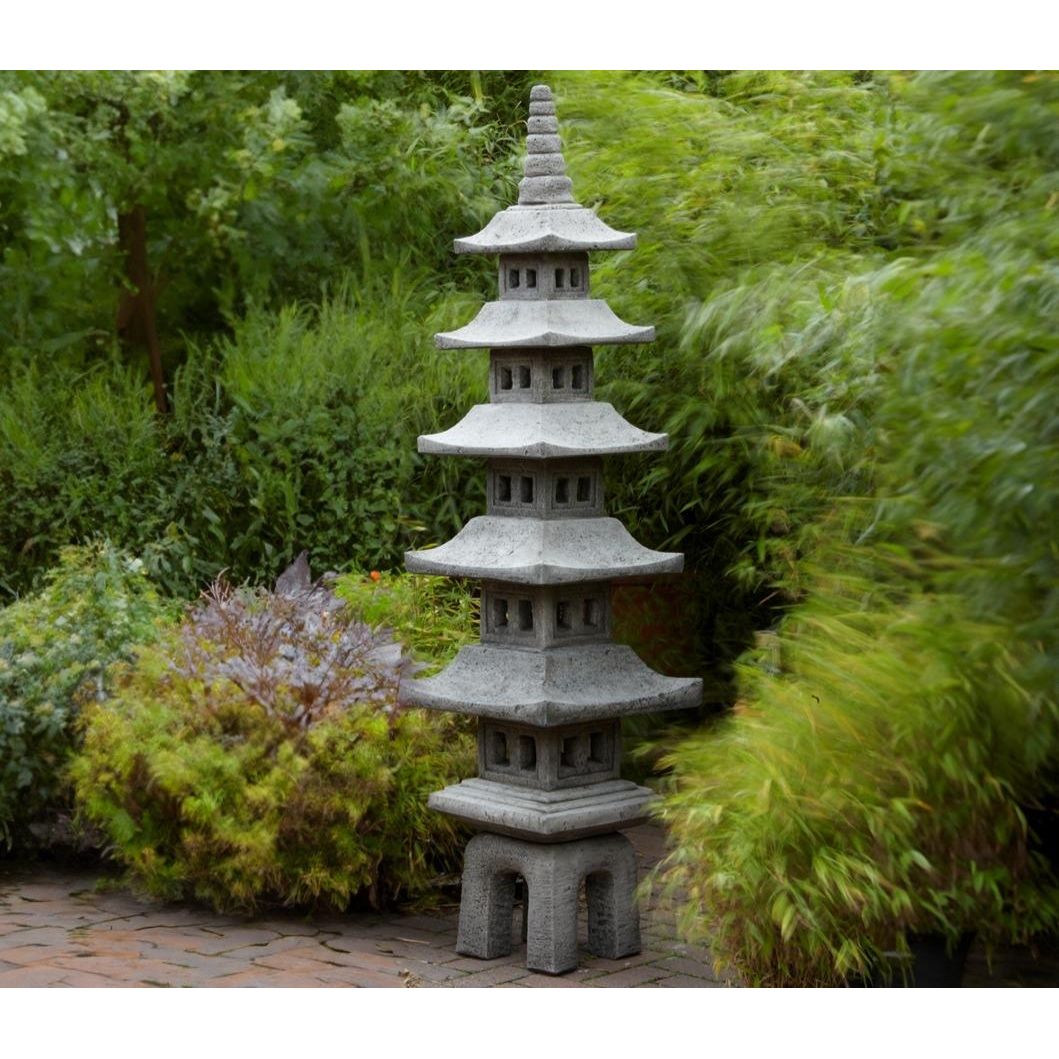A stone Japanese pagoda lantern is displayed in a lush garden setting, surrounded by green foliage and brick paving, enhancing a serene outdoor atmosphere.