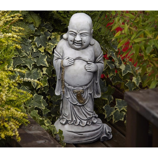 Grey smiling Buddha garden statue surrounded by green leafy plants on a wooden deck, adding a serene and spiritual touch to outdoor spaces with natural charm.