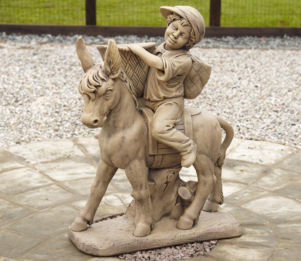 Stone garden ornament of a child with a hat riding a donkey, holding baskets. Set on a circular stone base with gravel and grass in the background.