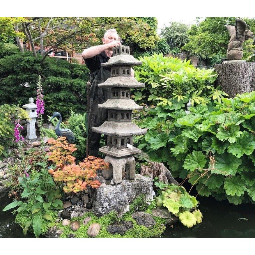 Man placing a decorative stone pagoda in a lush garden beside a tranquil pond, surrounded by vibrant green foliage and flowering plants, adding a serene ambiance.