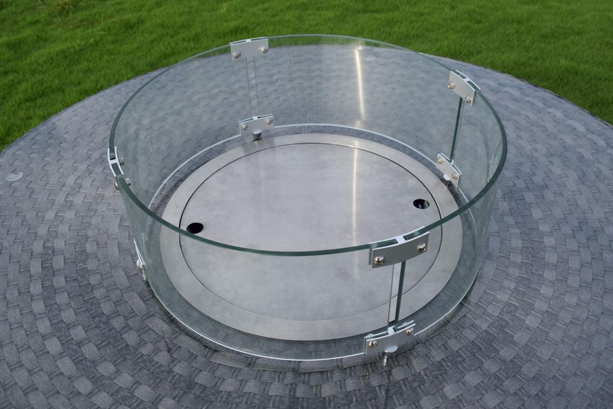 Round glass wind guard for an outdoor fire pit, featuring stainless-steel brackets on a rattan-patterned table surface, designed to protect flames from wind.