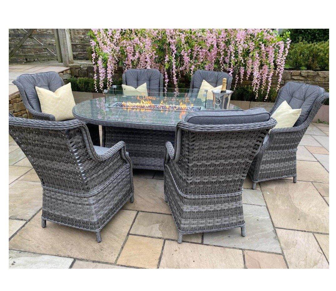 Grey rattan garden dining set with six cushioned chairs and a glass-top firepit table on a stone patio, surrounded by pink flowering plants for elegant outdoor dining.