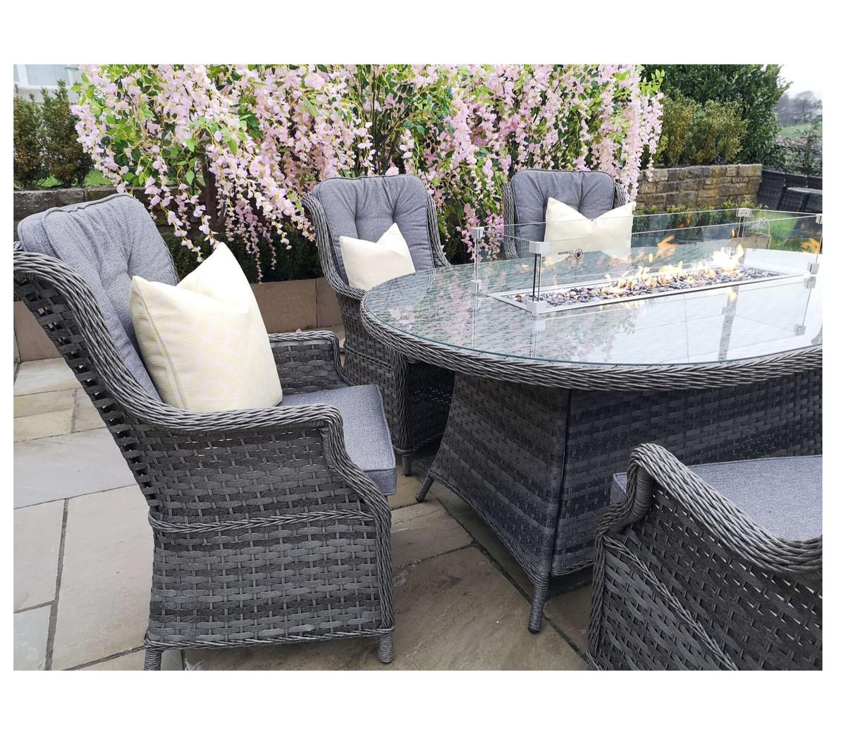 Grey rattan garden dining set with cushioned chairs and a glass table featuring a central gas fire pit. Surrounded by blooming pink flowers on a stone patio.