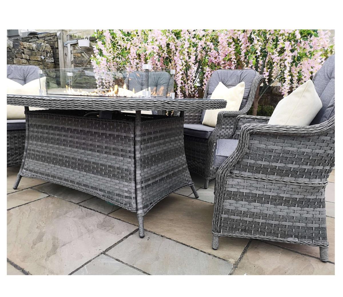 Grey wicker garden dining set featuring a rectangular table and matching armchairs. The scene is enhanced by decorative cushions and pink blossoms in the background.