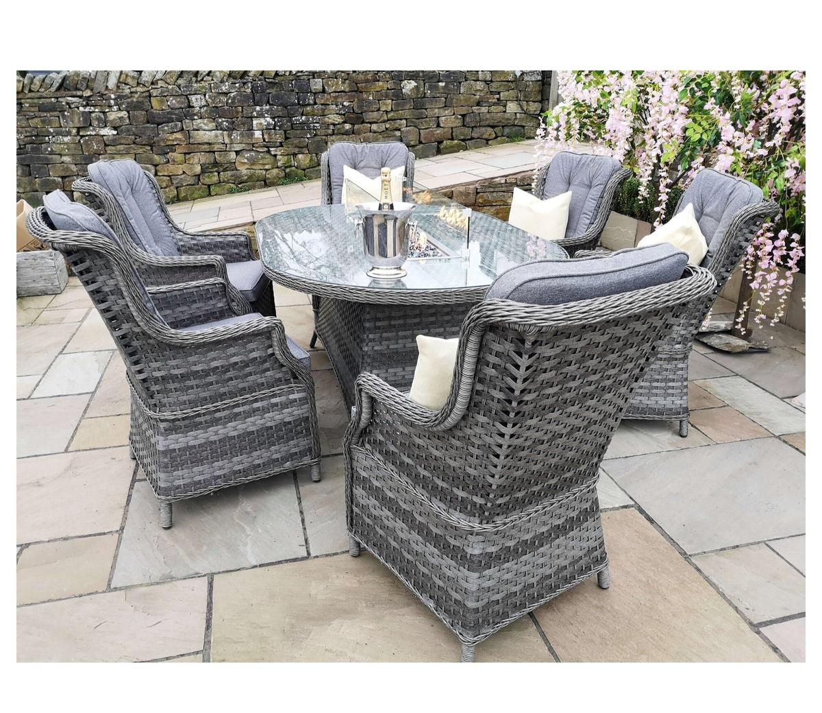 Outdoor patio set featuring a round glass table and comfortable grey wicker chairs with cushions, set on stone tiles. The area is decorated with blooming flowers and ambient lighting.
