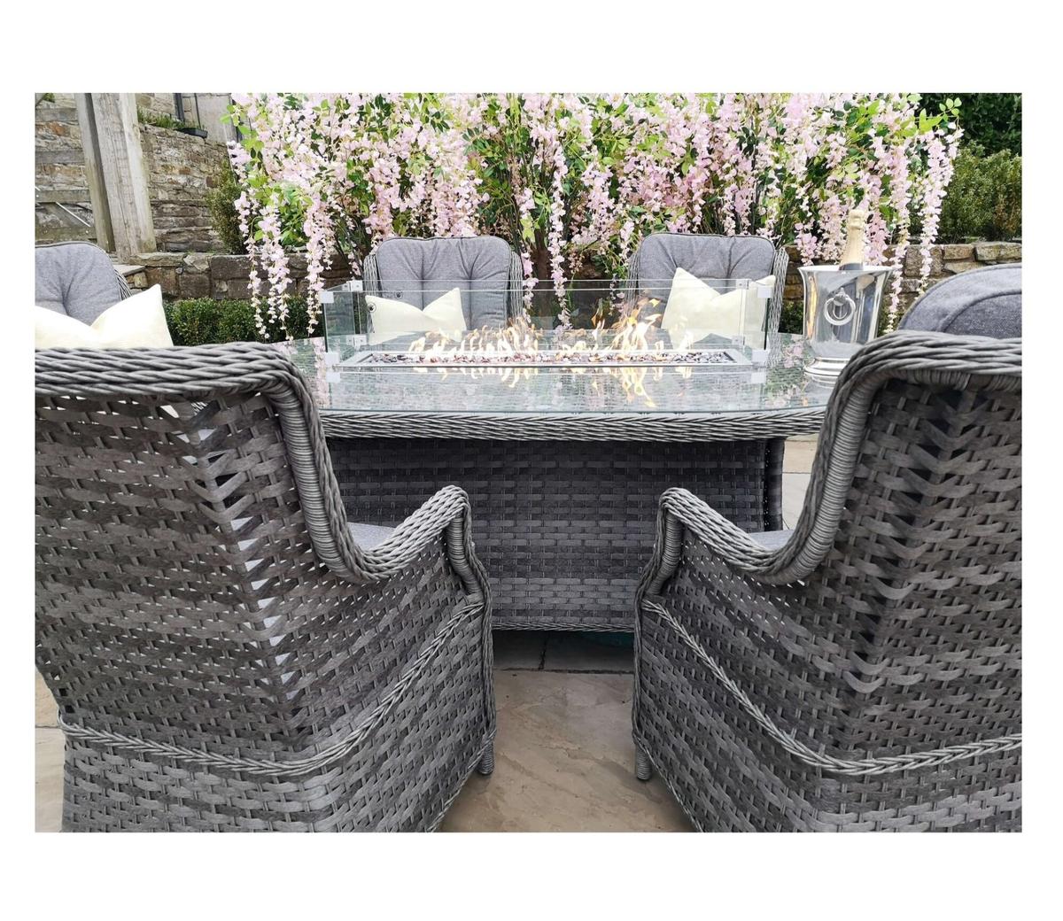 Grey rattan garden furniture set featuring a glass-top table with built-in fire feature, surrounded by cushioned chairs, placed in a patio setting with floral backdrop.