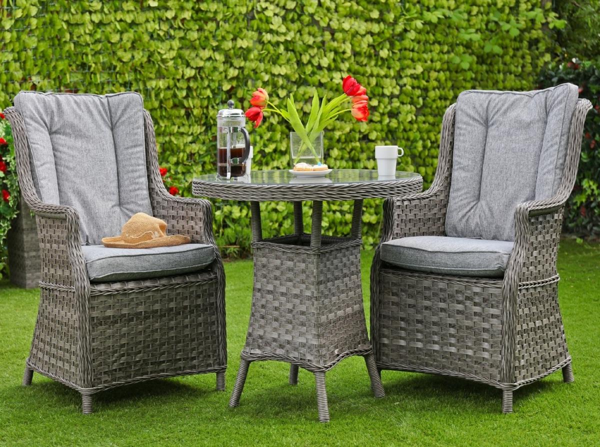 Outdoor wicker dining set featuring two grey cushioned chairs and a round table with a glass top. Decorated with red flowers, a coffee press, and a mug on a lush garden lawn.