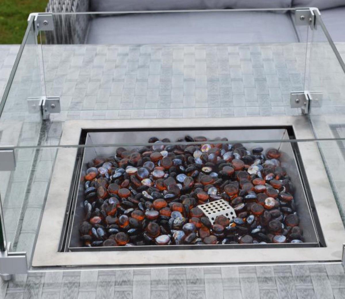 Modern outdoor fire pit with black and orange decorative stones enclosed in a glass panel, set on a textured grey surface, ideal for patio gatherings.