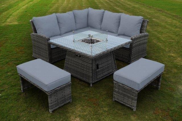 Outdoor grey rattan garden furniture set with plush cushions, arranged on grass. Features a corner sofa and glass-top fire pit table, framed by two cushioned benches.