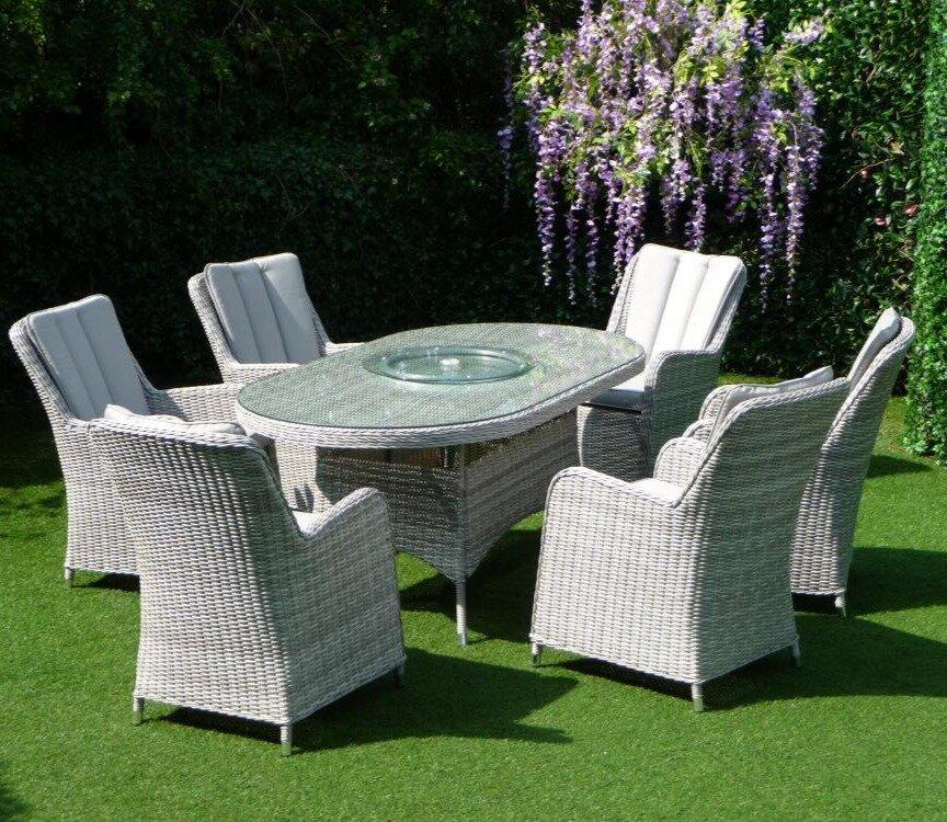 Six-piece grey rattan patio furniture set with cushioned chairs and an oval glass-top table, set on lush green grass, surrounded by vibrant purple wisteria.