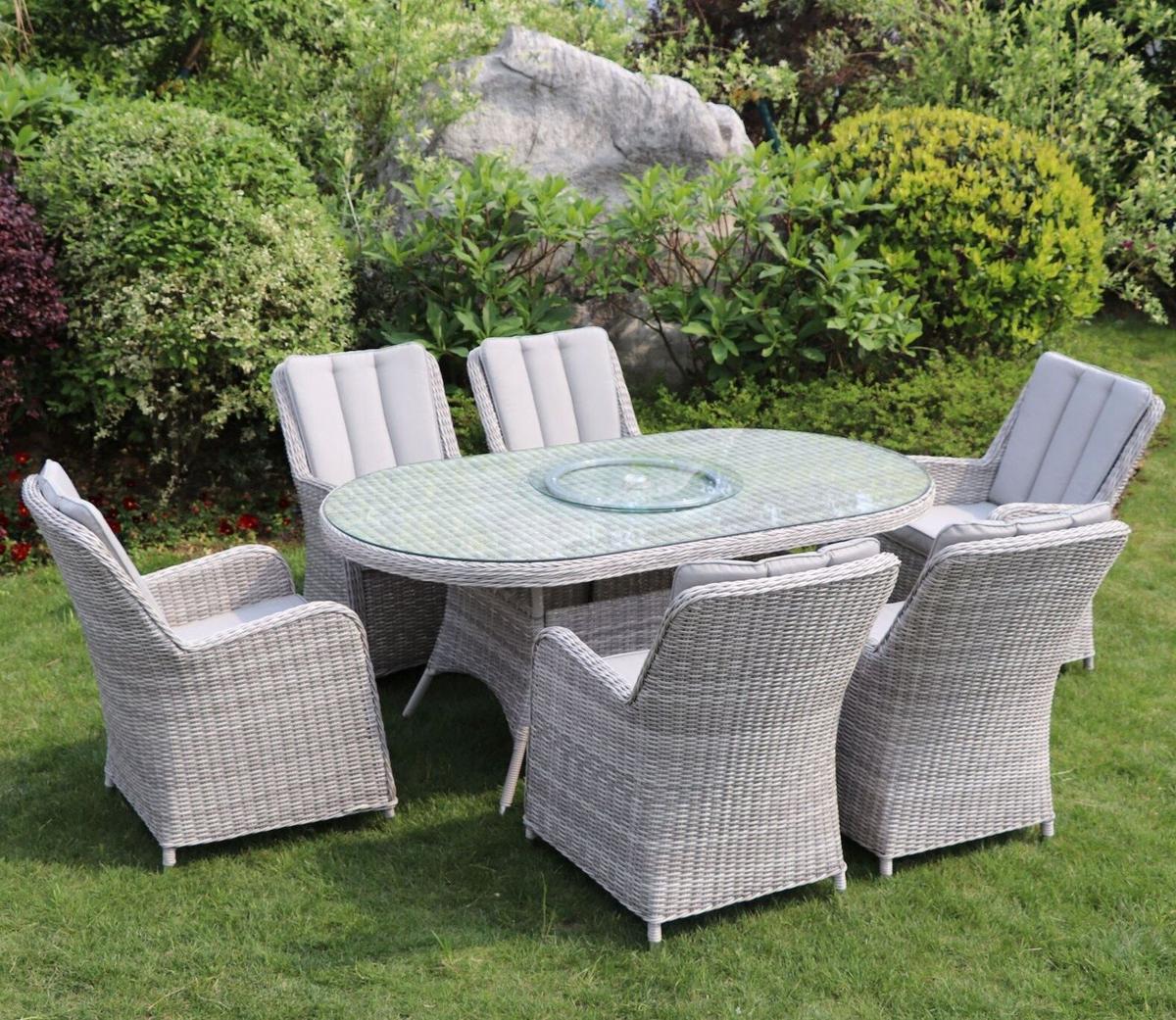 Outdoor garden set with six cushioned wicker chairs and an oval glass table in a lush green garden. Ideal for alfresco dining and relaxation in style.