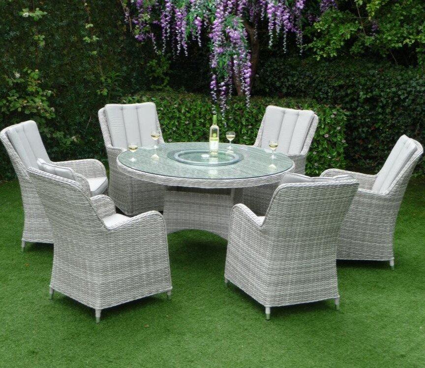 Outdoor garden set featuring a round glass-top table and six grey wicker chairs with cushions on a lush green lawn, wine and glasses on the table under hanging purple flowers.