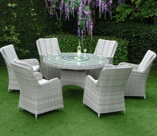 Outdoor garden set featuring a round glass-top table and six grey wicker chairs with cushions on a lush green lawn, wine and glasses on the table under hanging purple flowers.