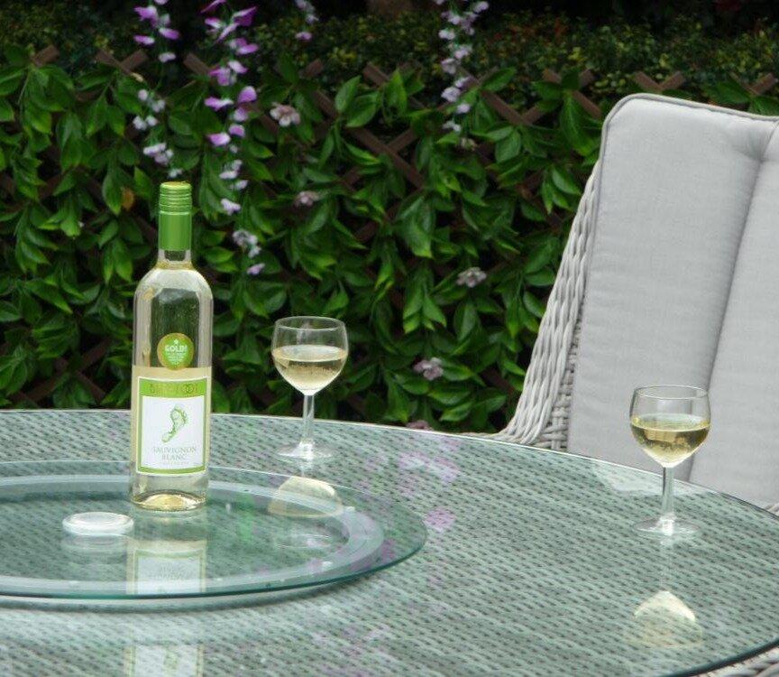 Bottle of Sauvignon Blanc wine on a glass table set outdoors, surrounded by lush greenery. Two wine glasses are partially filled, creating a serene garden ambience.
