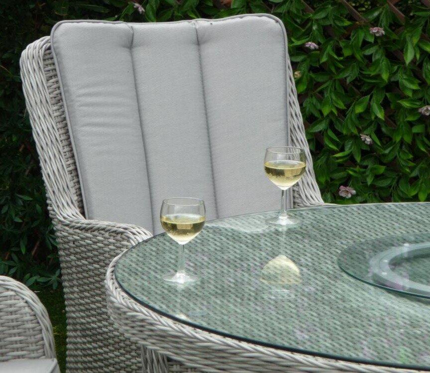 Outdoor rattan dining set featuring a glass-topped table and high-backed armchairs with cushioned seats. Two glasses of white wine add an elegant touch.