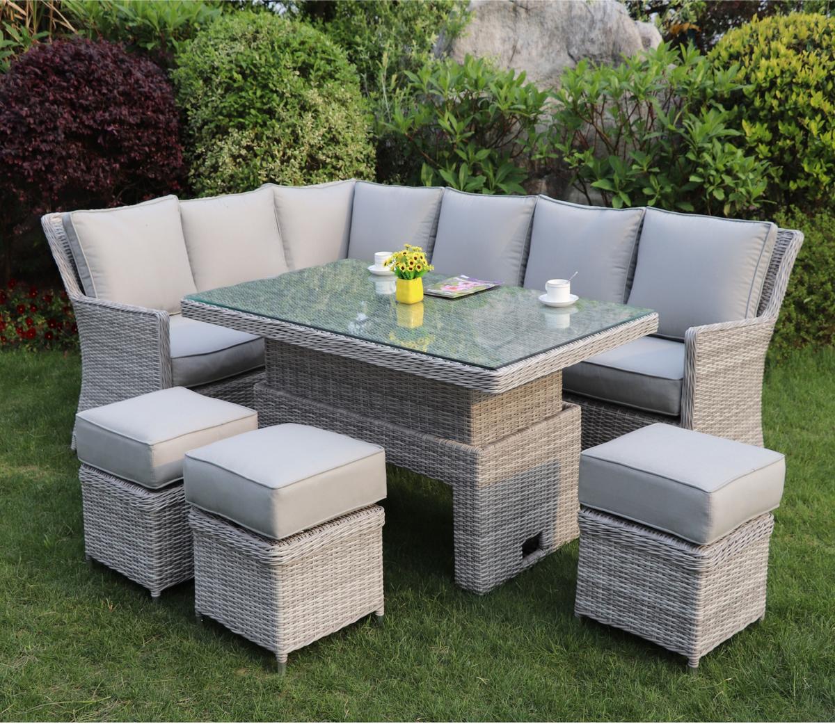 Grey wicker outdoor patio set featuring a corner sofa, glass-top dining table, and cushioned stools, surrounded by a lush garden landscape.