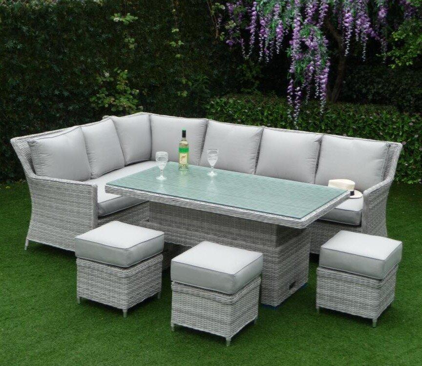 Grey rattan outdoor garden furniture set featuring an L-shaped sofa with cushions, a glass-topped rectangular table, and four cushioned stools on green lawn with purple flowers.