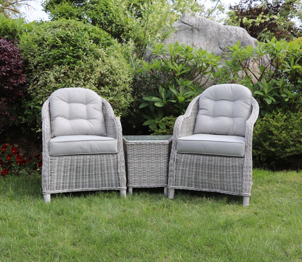 Grey wicker garden furniture set with two cushioned armchairs and a glass-topped side table, displayed on lush green grass with a backdrop of vibrant bushes.