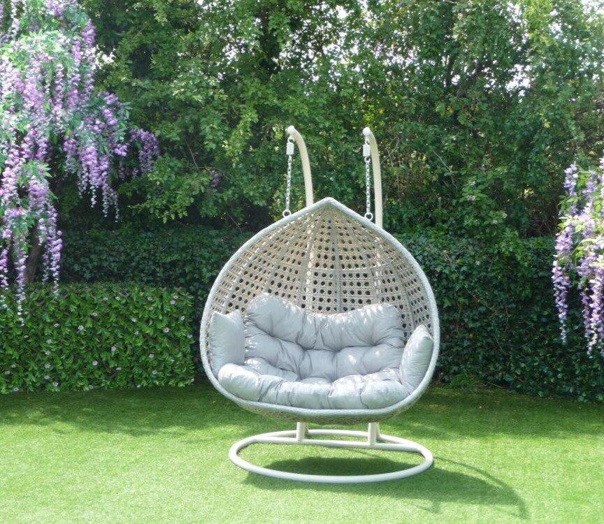 Elegant egg-shaped garden swing chair with grey cushion and woven frame, set on lush green grass with vibrant wisteria flowers and dense greenery in the background.