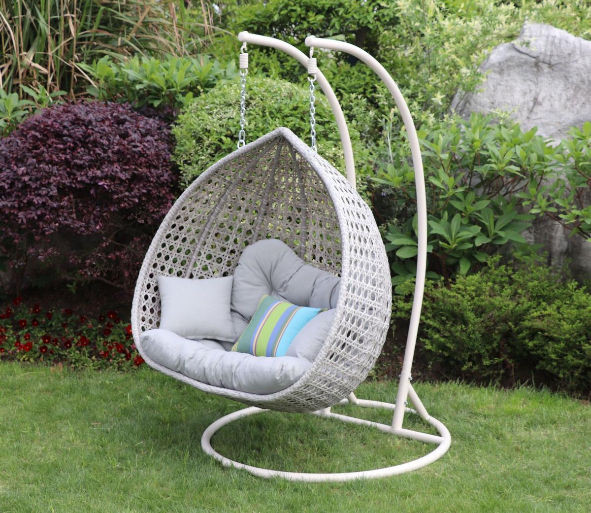 Hanging egg chair with a grey cushion in a garden setting. Features a metal stand and includes a colourful striped pillow. Surrounded by lush green foliage and flowers.