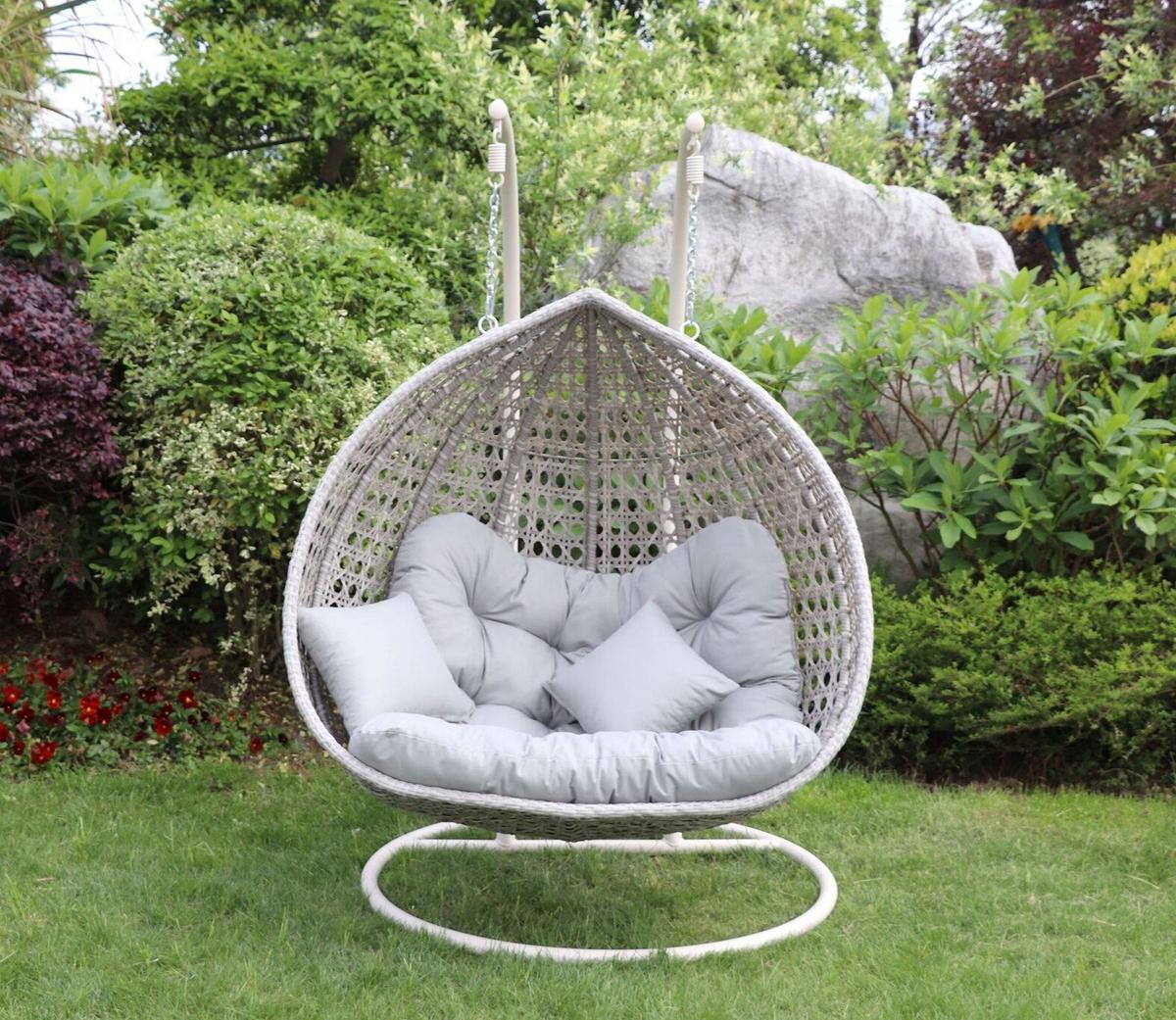 Cozy rattan egg chair with soft grey cushions for two, suitable for garden or patio, surrounded by lush greenery and flowers, perfect for outdoor relaxation.