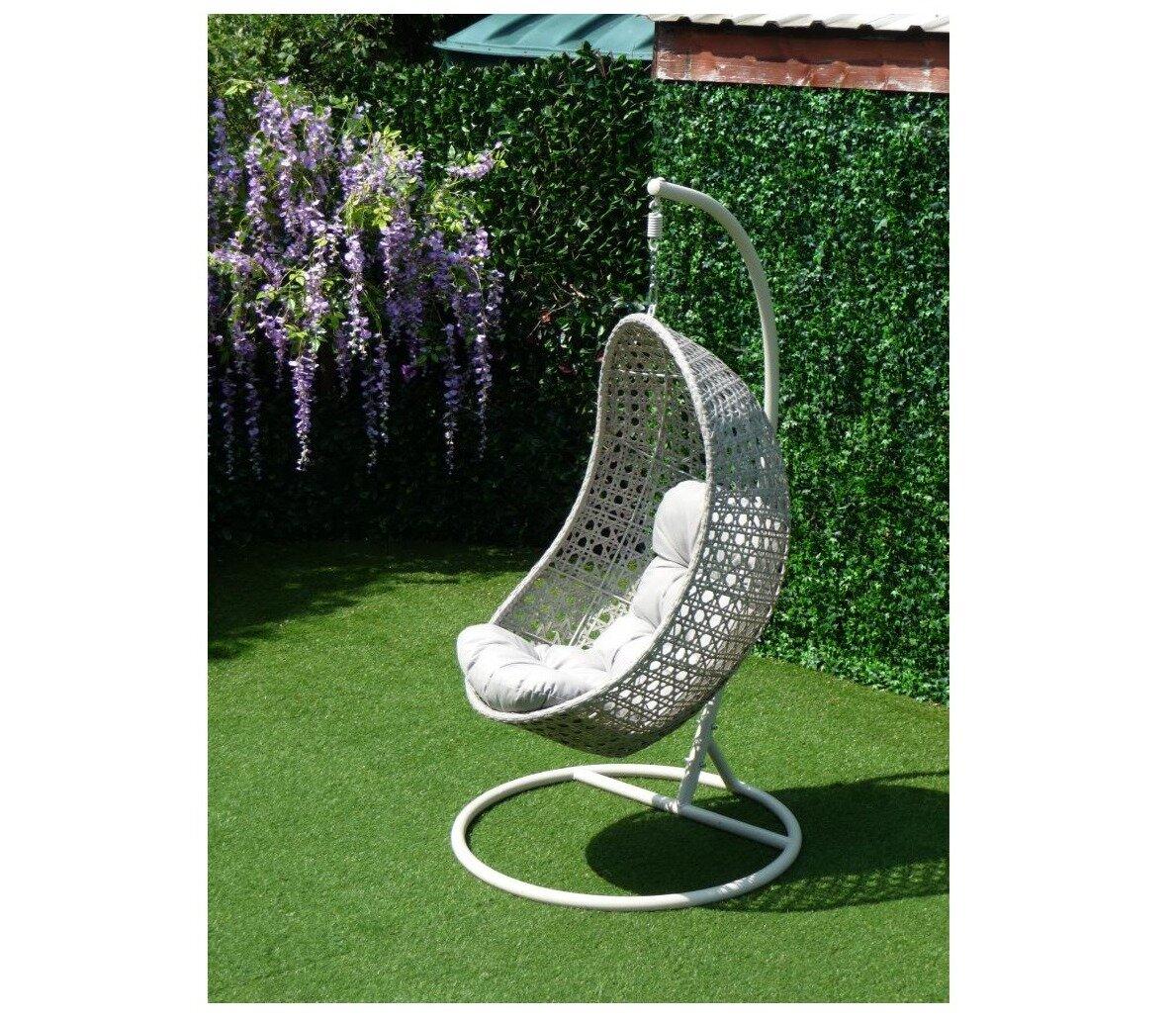 Hanging egg chair with a grey wicker frame and plush cushion, set on a stand. Positioned in a lush garden setting with artificial grass and floral backdrop.