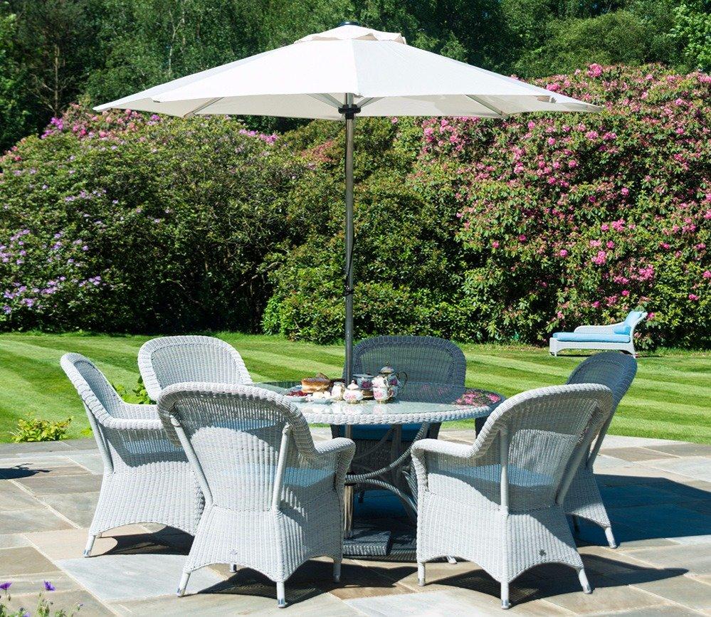 Outdoor patio setting featuring six grey wicker chairs around a round table with a white parasol, situated on a stone terrace amidst lush garden greenery.