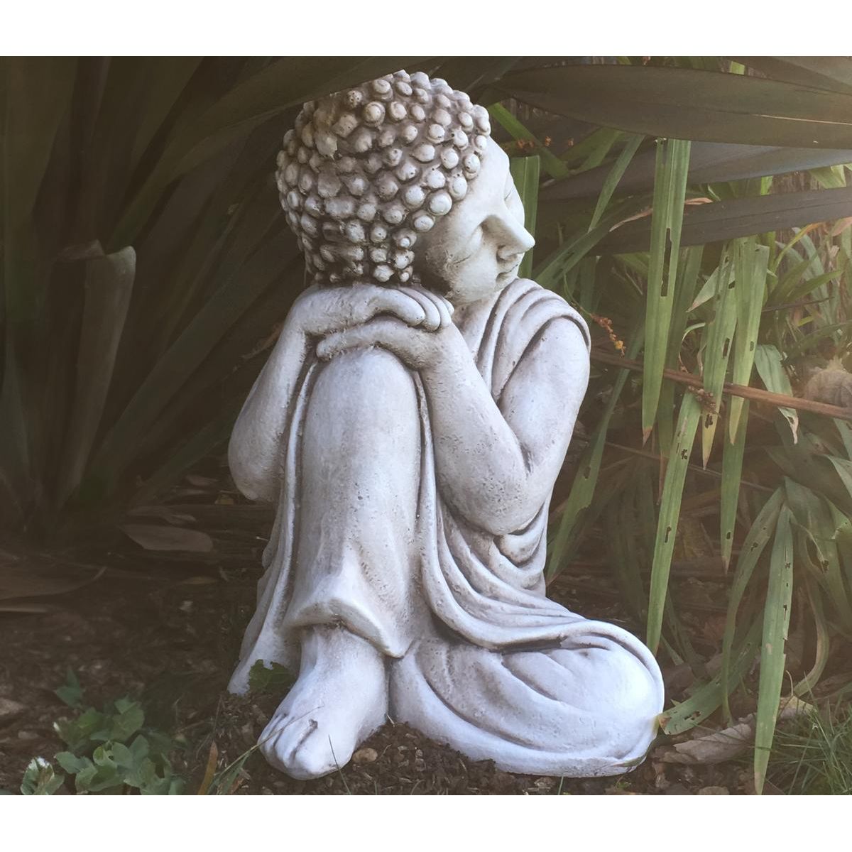 Stone statue of a serene figure sitting in a contemplative pose, surrounded by lush green foliage. Ideal for adding a peaceful touch to garden decor.