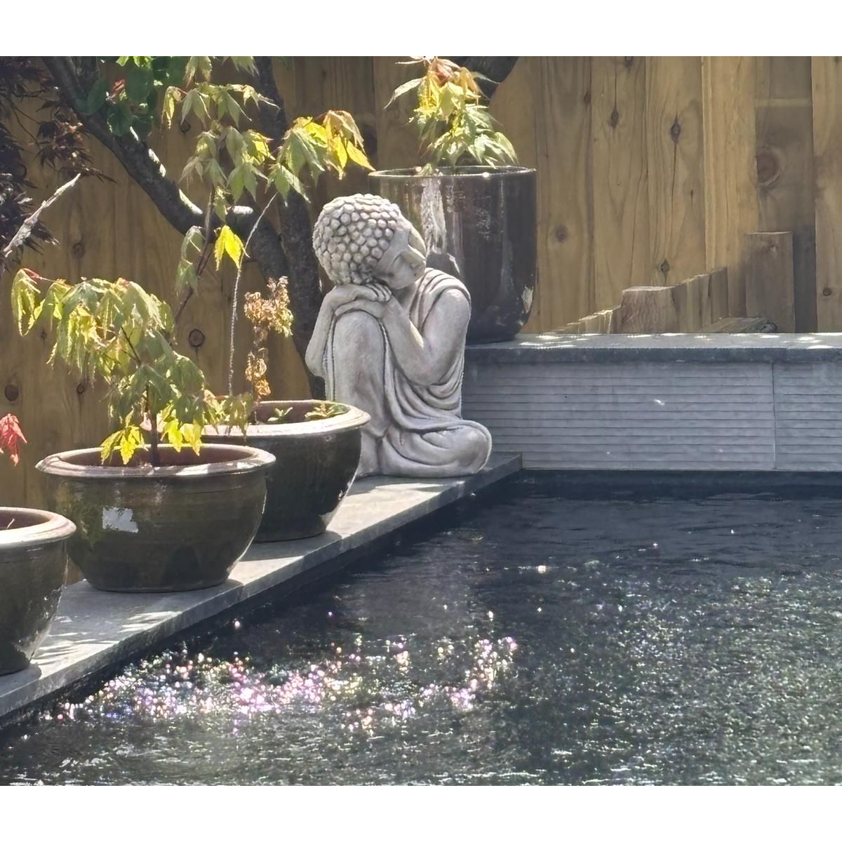 Serene garden scene featuring a contemplative Buddha statue surrounded by lush potted plants, set beside a tranquil stone pond reflecting sunlight.
