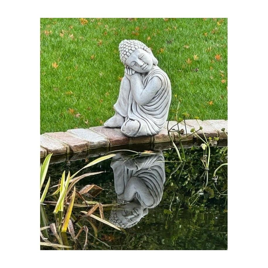 A serene garden statue of a meditating figure sits beside a reflective pond, surrounded by lush grass. The peaceful scene adds tranquility to outdoor spaces.
