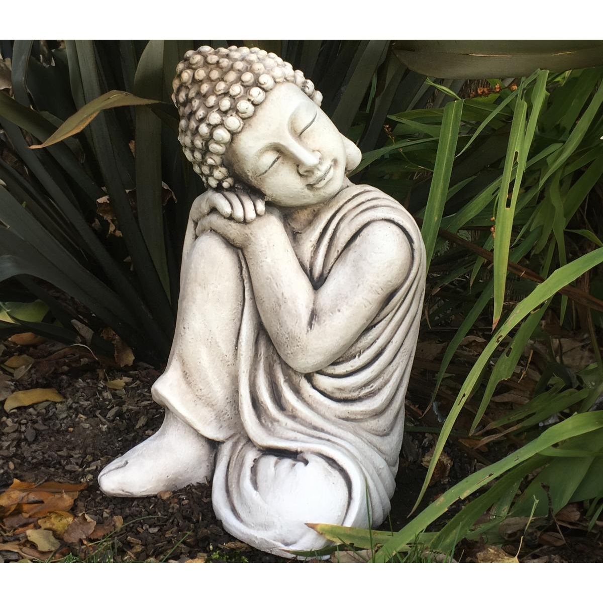A serene stone Buddha statue sits in a peaceful garden with lush green plants. The statue features a meditative pose and intricate details on its robe and hair.