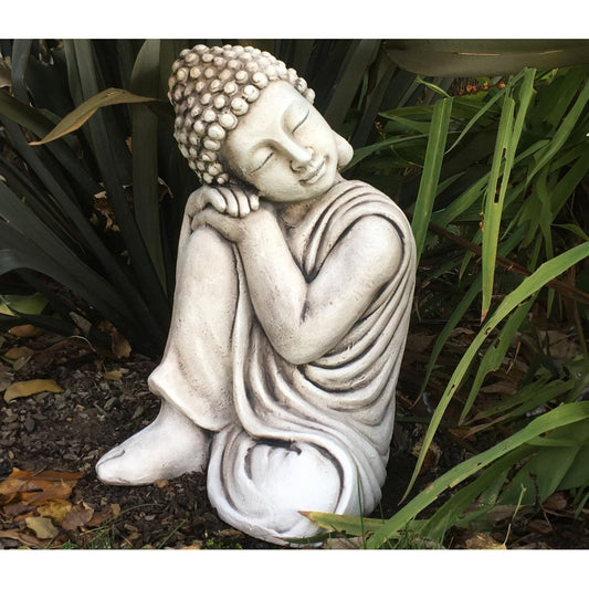 A serene stone Buddha statue sits in a peaceful garden with lush green plants. The statue features a meditative pose and intricate details on its robe and hair.