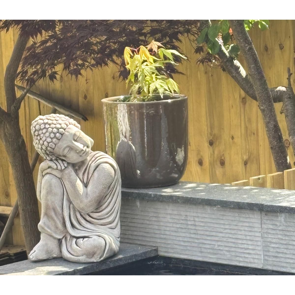 Tranquil outdoor scene featuring a serene Buddha statue resting against a potted plant, set on a grey stone platform with wooden fence backdrop.
