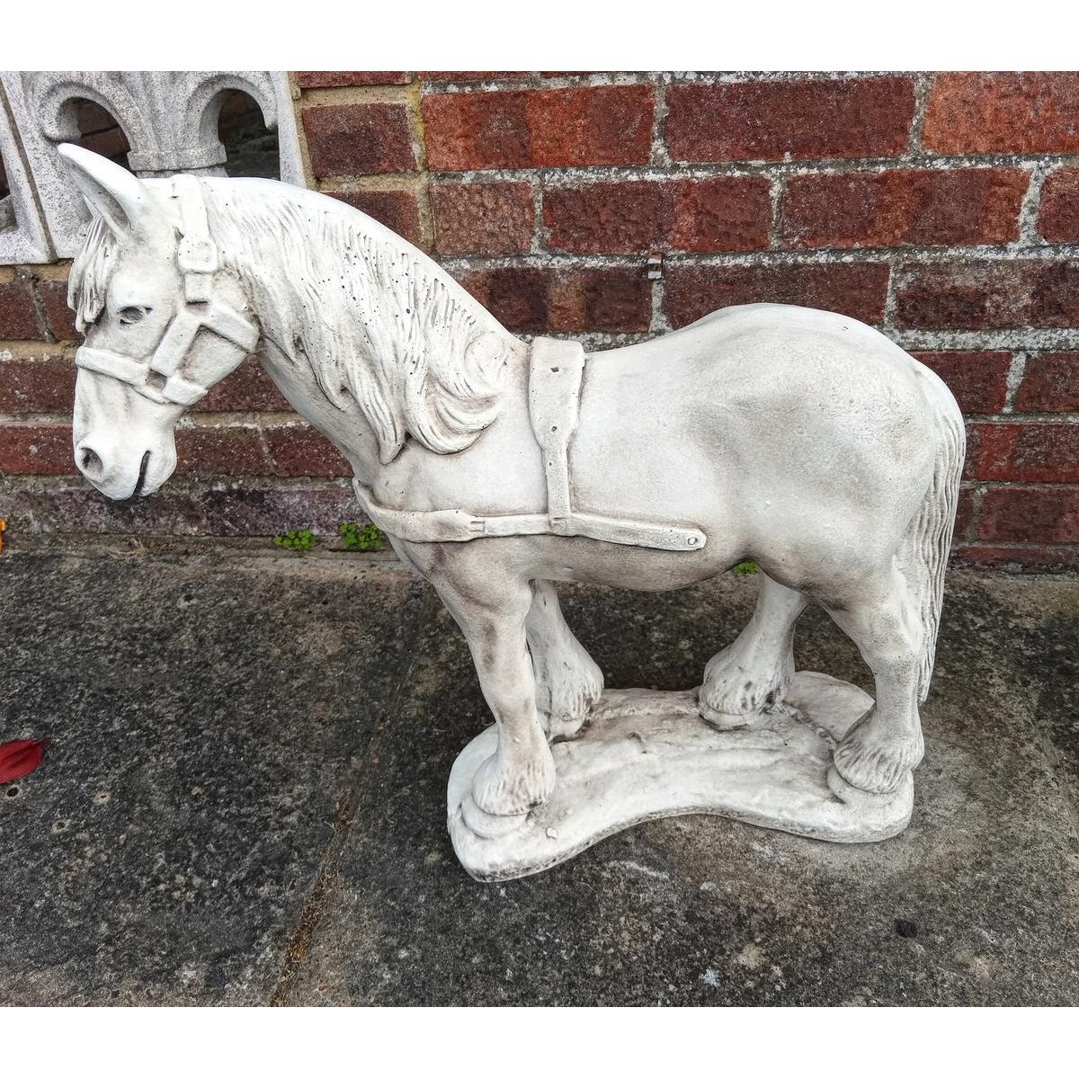 Statue of a horse made from stone with a detailed harness, standing on a textured platform against a brick wall backdrop, showcasing fine craftsmanship.