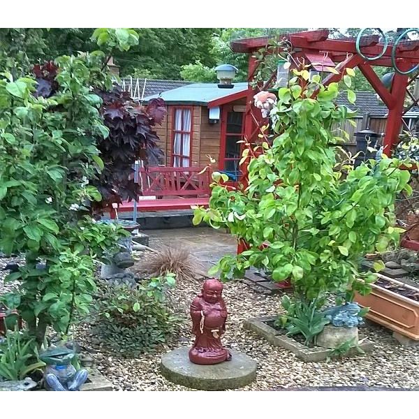 Lush garden with diverse plants and a red Buddha statue on gravel. In the background, a charming wooden building with a red pergola and vibrant foliage.
